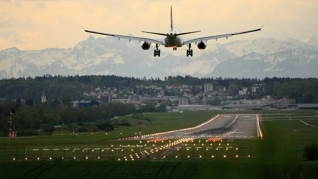 Ein zweistrahliges Verkehrsflugzeug ist von hinten im Landeanflug auf den Flughafen Zürich zu sehen. Dabei ist die beleuchtete Landebahn und im Hintergrund Berge zu sehen.