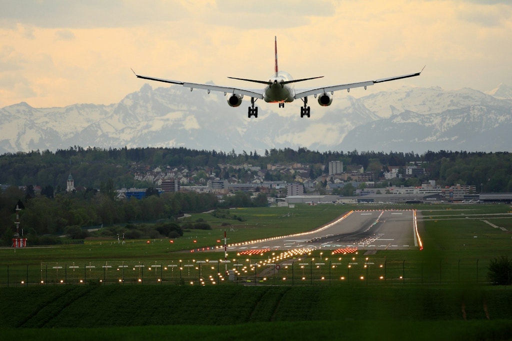 Ein zweistrahliges Verkehrsflugzeug ist von hinten im Landeanflug auf den Flughafen Zürich zu sehen. Dabei ist die beleuchtete Landebahn und im Hintergrund Berge zu sehen.